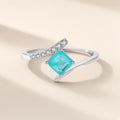 Bague Calanque argent 925, pierre d'imitation carrée bleu-vert posée de biais sur un anneau croisé, sur fond crème