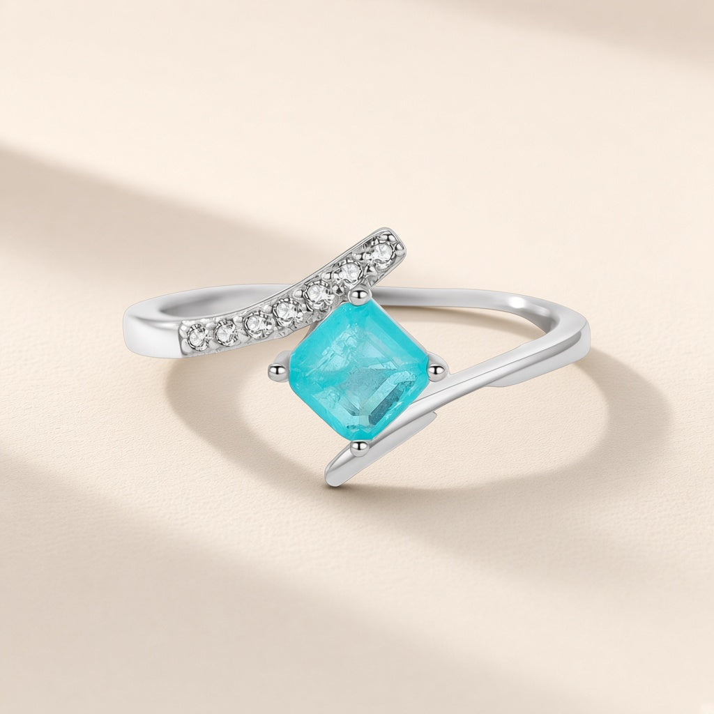 Bague Calanque argent 925, pierre d'imitation carrée bleu-vert posée de biais sur un anneau croisé, sur fond crème