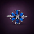 Bague Bleuet argent 925, six zirconiums cubiques bleu roi taillés en goutte autour d'une petite pierre ronde claire, sur fond aubergine