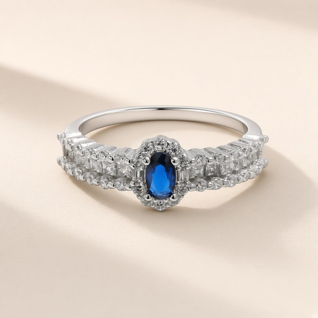 Bague Minuit argent 925, zirconium cubique ovale bleu roi entouré de petites pierres claires, anneau large serti, sur fond crème